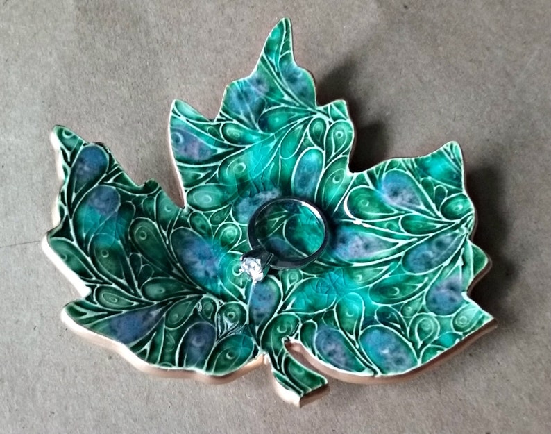 May include: A ceramic leaf-shaped dish with a green and blue swirling pattern. The dish has a gold rim and a ring is resting in the center.