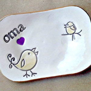 May include: White ceramic dish with a gold rim. The dish has a hand-painted design of two birds and a heart with the word "oma" written on it.