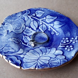 May include: A blue and white ceramic ring dish with a floral design. The dish has a gold rim and a ring is sitting in the center.