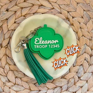 May include: A green clover-shaped keychain with the name "Eleanor" and "TROOP 12345" in white lettering. It has a green tassel and silver hardware. The keychain rests on a light-colored, round dish.
