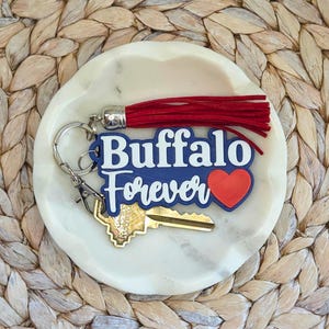 May include: A keychain with the words "Buffalo Forever" in white script on a blue background, with a red heart. It has a red tassel and a gold key attached. The keychain rests on a white marble dish.