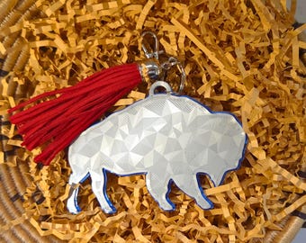 Op de afbeelding: Een zilverkleurige sleutelhanger met een geometrisch patroon en blauwe rand, bevestigd aan een rode kwast. De sleutelhanger wordt gepresenteerd op geel gescheurd papier in een geweven mand.