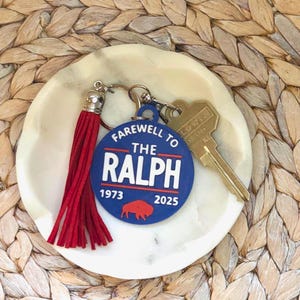 May include: A blue and white keychain with the text "Farewell to the Ralph" and the years 1973 and 2025. A red tassel and a gold key are attached. The keychain is on a white marble dish.