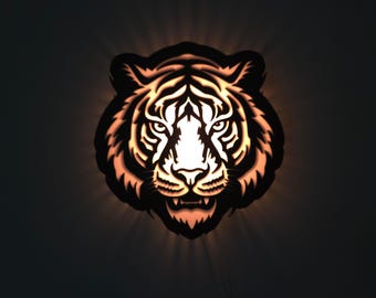 Op de afbeelding: Een decoratief wandkunstwerk met een silhouet van een tijgerkop. Het ontwerp is uit een donker materiaal gesneden, met de details en ogen van binnenuit verlicht door een warme, oranje gloed. Het gezicht van de tijger is gecentreerd, met gedetailleerde vachtpatronen.