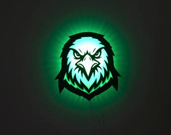 Op de afbeelding: Een gloeiend adelaarskop-silhouet met een felle uitdrukking, verlicht door groen licht. De kenmerken van de adelaar zijn scherp gedefinieerd, met een zwarte omtrek en een helder, wit en groen interieur. Het ontwerp is op een donkere achtergrond gemonteerd.