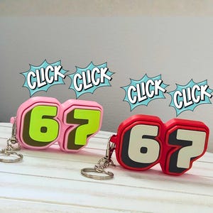 May include: Two keychains shaped like the number 67. One is pink with green numbers, the other is red with white numbers. Each keychain has a silver ring. Comic book style "CLICK" speech bubbles are above the keychains.