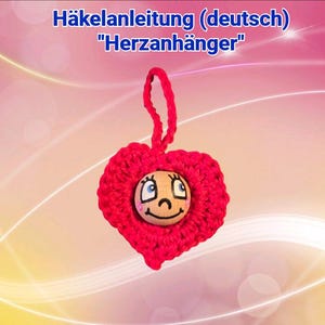 May include: A handmade red crocheted heart-shaped ornament with a smiling wooden face. The ornament has a loop for hanging and is set against a gradient pink and yellow background. Text at the top reads "Häkelanleitung (deutsch) "Herzanhänger".