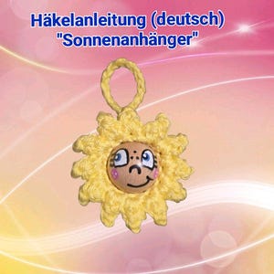 May include: A handmade yellow crochet sunflower with a smiling face, featuring blue eyes and pink cheeks. The sunflower has a loop for hanging. Text at the top reads "Häkelanleitung (deutsch) Sonnenanhänger".
