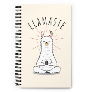 Op de afbeelding: Een spiraalgebonden notitieboek met een crèmekleurige kaft. De kaft toont een cartoon lama in een yogahouding, met de tekst "LLAMASTE" erboven. De lama is wit met roze accenten. Zwarte spiraalbinding.
