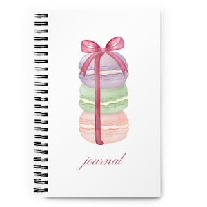 Könnte beinhalten: Ein Stapel aus drei pastellfarbenen Macarons, die mit einem rosa Band zusammengebunden sind. Die Macarons sind von unten nach oben gestapelt: rosa, grün und lila. Der Text "Journal" ist in Kursivschrift unter den Macarons geschrieben.