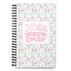 Puede incluir: Un cuaderno de espiral con un estampado floral en colores pastel. La portada presenta el texto "It's Okay not to be Okay" en letras rosas sobre un fondo blanco. La encuadernación es en espiral negra.