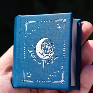 Op de afbeelding: Een klein, blauw boek met zilveren hemelse ontwerpen en de tekst "Written in the Stars". De omslag heeft een maan en kristal ontwerp. Het boek wordt in een hand gehouden.