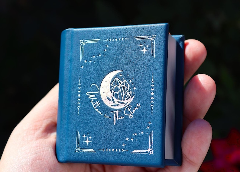 Peut inclure: Un petit journal reli&eacute; en cuir bleu avec un motif en argent gaufr&eacute; repr&eacute;sentant une lune croissante, un cristal et les mots "Written in the Stars".
