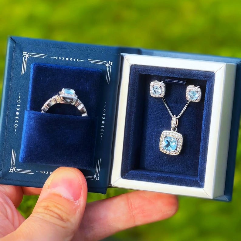 Peut inclure: Un coffret &agrave; bijoux en velours bleu contenant une bague, des boucles d'oreilles et un collier. Les bijoux sont sertis de pierres bleues et d'argent.