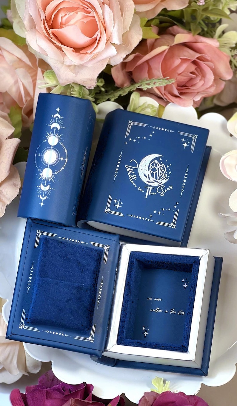 Peut inclure: Une bo&icirc;te &agrave; bague en velours bleu avec un motif de lune et d'&eacute;toiles. La bo&icirc;te est ouverte et l'int&eacute;rieur est doubl&eacute; de velours bleu. Le texte "Written in the Stars" est imprim&eacute; sur la bo&icirc;te.