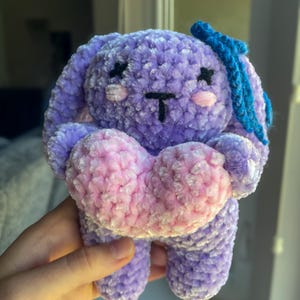 May include: A handmade plush bunny in shades of purple, holding a pink and white heart. The bunny has black stitched eyes and a black stitched mouth. A blue bow is attached to the top of its head. The plush toy is soft and cuddly.
