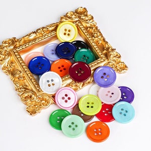 May include: A selection of colourful, round buttons in shades like yellow, blue, green, and red. The buttons are arranged in and around a decorative gold-coloured frame. Each button has four holes for sewing.