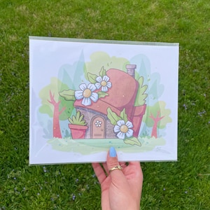 Puede incluir: Una ilustración caprichosa de una pequeña casa con un techo marrón y una puerta blanca. La casa está rodeada de hojas verdes y flores blancas. La ilustración está hecha en un estilo de dibujos animados.