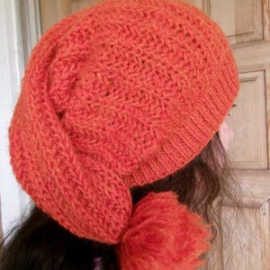 Op de afbeelding: Een feloranje gebreide beanie met een pluche pompon. De beanie is gemaakt met een textuurpatroon.