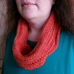 May include: An orange knitted infinity scarf. The scarf is made of a thick yarn and has a textured appearance. The scarf is wrapped around the neck and is set against a teal green top.