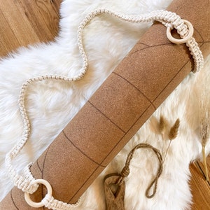 Könnte beinhalten: Eine braune Kork-Yogamatte mit einem weißen gehäkelten Riemen und Holzringen. Die Matte liegt auf einer weißen, flauschigen Oberfläche.