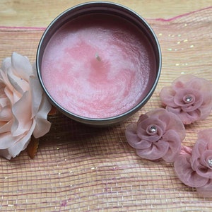 May include: A pink candle in a silver tin with a white wick, surrounded by three pink fabric flowers with rhinestones in the center. The flowers and candle are on a pink and brown woven surface.