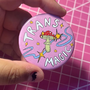 Puede incluir: Botón rosa con el texto "TRANS MAGIC" en blanco, un hongo de dibujos animados con un sombrero rojo y puntos blancos, y un remolino azul con estrellas amarillas.