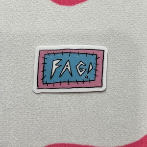 Könnte beinhalten: Ein Aufkleber mit einem rosa und weißen Rand und einer blauen Mitte. Die Mitte hat einen gestickten Rand und das Wort "FAG!" in Schwarz geschrieben.