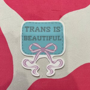 May include: A blue and white sticker with the text "TRANS IS BEAUTIFUL" in a stitched font. A pink and white bow with a heart in the center is below the text.