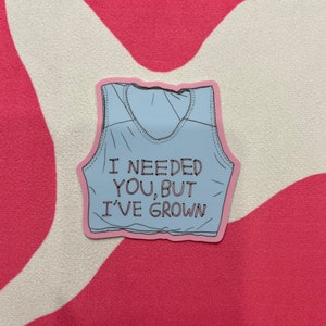 Puede incluir: Un top de tirantes azul claro con ribetes rosas y el texto "I NEEDED YOU, BUT I'VE GROWN" impreso en la parte delantera.