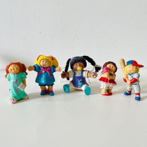 Peut inclure: Ensemble de cinq figurines Cabbage Patch Kids. Les poupées sont dans diverses poses et tenues, dont un joueur de baseball, une fille avec une glace et un bébé avec une sucette. Les poupées sont aux couleurs vives et ont des détails.