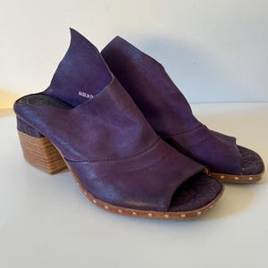 May include: A pair of open-toe, purple leather mules with a stacked wooden heel. The shoes have a unique design with a folded-over upper and are embellished with small gold-colored studs around the sole.