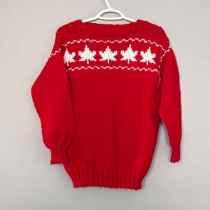 May include: A red knitted sweater featuring a row of white leaf patterns across the chest. The sweater has long sleeves and is displayed on a white hanger. The background is a plain grey.