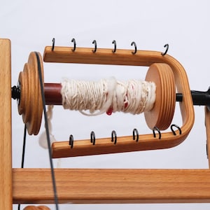 May include: A wooden spinning wheel with a light brown finish. The wheel has a central spindle wrapped with white yarn, and a series of black hooks. The frame is made of light-coloured wood.