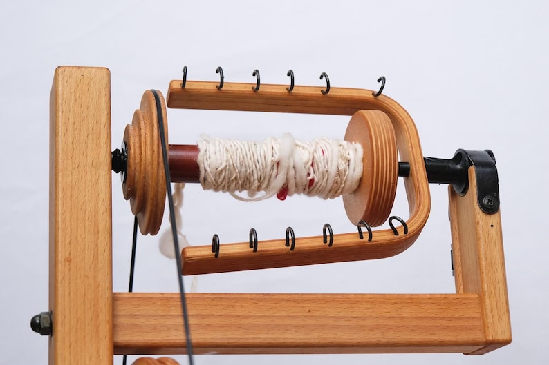 May include: A wooden spinning wheel with a light brown finish. The wheel has a central spindle wrapped with white yarn, and a series of black hooks. The frame is made of light-coloured wood.