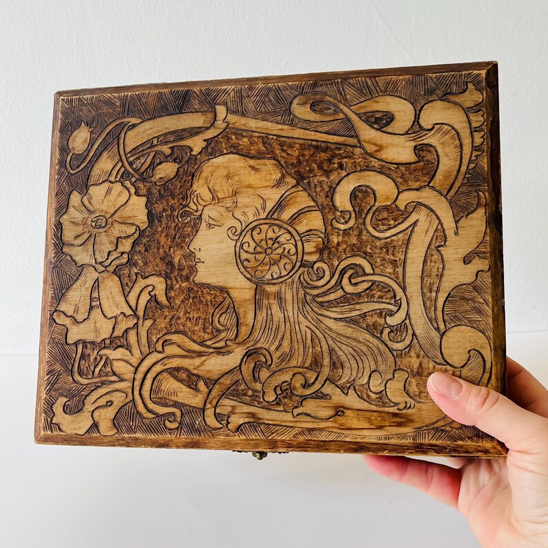 May include: Wooden rectangular box with a detailed pyrography design. The artwork features a woman's profile with flowing hair and floral elements. The box is stained a warm brown color, showcasing the wood grain and intricate carvings.
