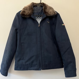 May include: A navy blue jacket with a brown faux fur collar. The jacket has a zip fastening and two pockets. The jacket has a small Italian flag patch on the right sleeve and a small airplane logo on the left chest.