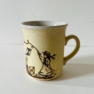May include: A yellow ceramic mug with a brown illustration of a girl flying a kite. The mug has a brown handle.