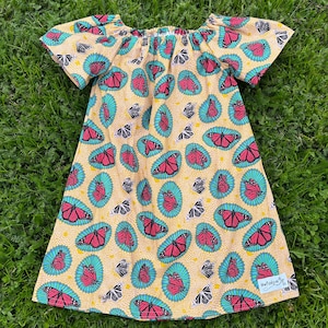 May include: A yellow dress with a butterfly pattern. The dress features short sleeves and a gathered neckline. The butterflies are pink and black, set against a teal and yellow background. The dress is made of a lightweight fabric.
