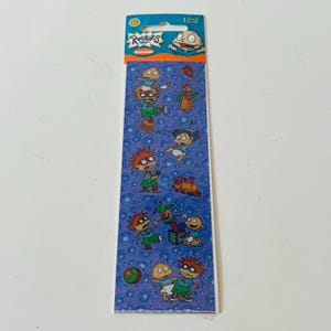 Puede incluir: Una hoja vertical de pegatinas de Rugrats con varios personajes de dibujos animados, incluidos Tommy Pickles y Carlitos Finster. El fondo es un patrón azul de círculos giratorios. El encabezado muestra el logotipo de Rugrats y la marca Nickelodeon.
