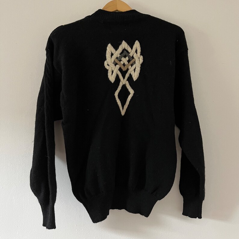 May include: Black sweater with a cream-colored embroidered design on the back. The design features a geometric pattern with diamond and knot-like shapes. The sweater has long sleeves and a crew neck.
