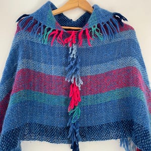May include: A blue and pink striped knitted poncho with a fringe trim. The poncho has a hood and is made of a soft, textured yarn.