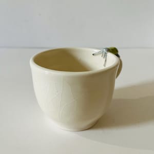 Peut inclure: Une tasse en céramique de couleur crème avec une finition brillante et un motif de glaçure craquelée visible. La tasse présente une petite figurine d'insecte sculptée en vert et gris fixée sur le bord supérieur, servant d'accent décoratif.