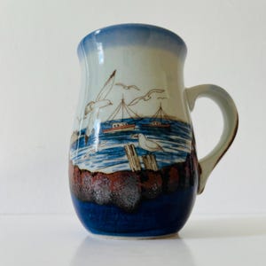Peut inclure: Mug en céramique avec une scène côtière. Le mug présente un dégradé bleu et blanc sur le dessus, une anse et une illustration détaillée de mouettes, de bateaux et de la mer. La base du mug est de couleur bleu foncé.