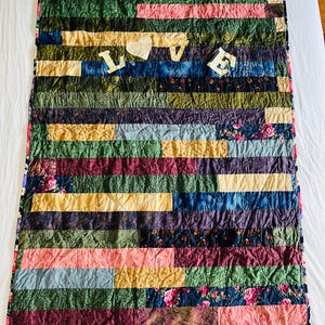 May include: A rectangular quilt featuring a patchwork of horizontal fabric strips in various colours including green, blue, pink, yellow, and dark floral patterns. The word LOVE is appliqued in white fabric letters across the upper centre section.