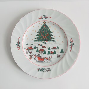 Puede incluir: Plato navideño de cerámica blanca con borde rojo y escena invernal festiva. El plato presenta un árbol de Navidad decorado, casas y un trineo tirado por caballos. Acentos verdes y rojos adornan el borde del plato.