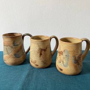 Może przedstawiać: Trzy ceramiczne kubki z brązową, beżową i niebieską glazurą. Kubki mają rustykalny, ręcznie robiony wygląd.