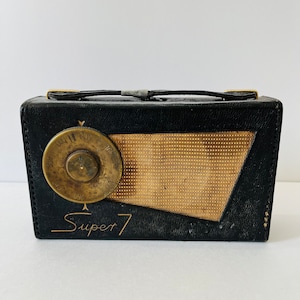 Peut inclure: Radio Super 7 noire vintage avec un couvercle de haut-parleur et un cadran de réglage dorés. La radio a une poignée et des accents dorés. Le texte "Super 7" est imprimé sur le devant.