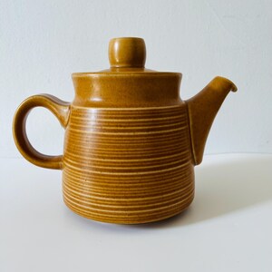 Puede incluir: Una tetera de cerámica color amarillo mostaza con cuerpo cilíndrico y detalles de estrías horizontales, asa curva, pico corto y tapa a juego con un pomo redondeado. La superficie presenta un acabado mate, liso y uniforme en todo el objeto.