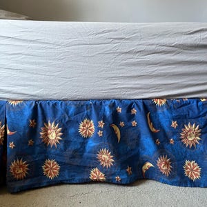 May include: A blue bed skirt featuring a repeating pattern of golden yellow suns with faces, crescent moons, and small stars. The fabric has a textured appearance and is positioned beneath a light gray bed sheet on a mattress base.